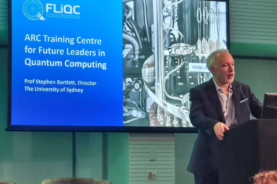 A photo of Prof Stephen Bartlett speaking at a lectern with a screen projection behind him of the FLiQC logo.
